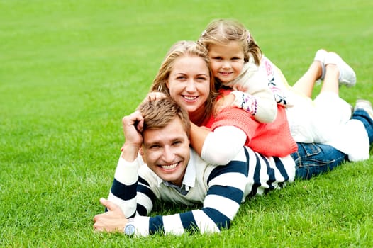 Smiling family of three piled on top of each other. All relaxing on lush green ground