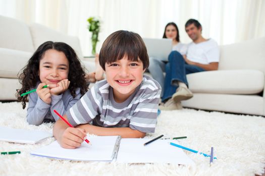 Cute children drawing lying on the floor with their parents in the background