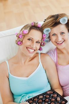 Delighted women wearing hair rollers eating chocolate looking at the camera in the living-room