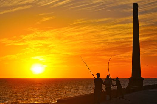 Silhouettes of three men on the coast fishing at sunset.