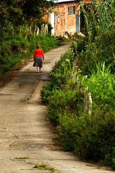 An elderly woman walks down a road in rural South America.