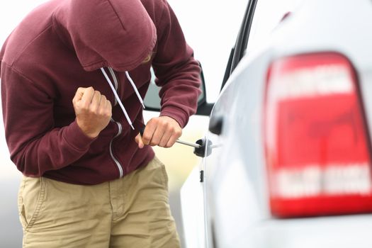 View down the side of a car to a man in a hooded top breaking into a car with a screwdriver in order to steal it