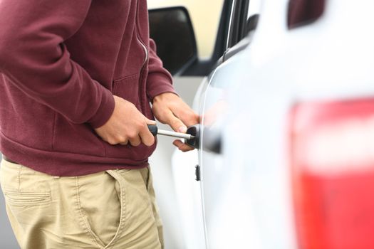 View down the length of a car to a man committting a car break in and theft with a screw driver inserted into the lock