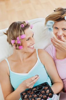 Beautiful female friends with hair rollers eating chocolate at home on a sofa