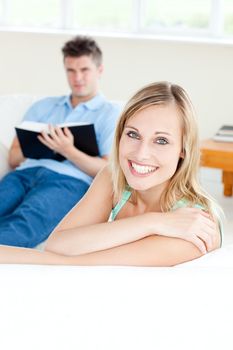 Girlfriend smiling at the camera while her boyfriend is reading a book on the sofa in the living-room