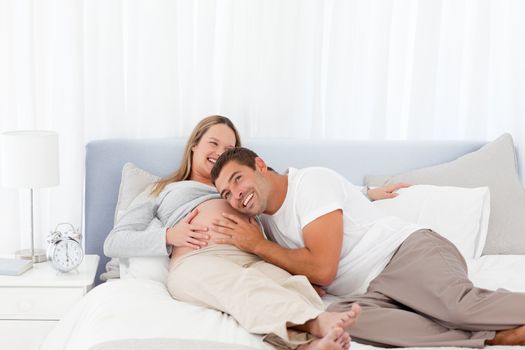 Funny man putting his head on the belly of his pregnant wife while relaxing on the bed