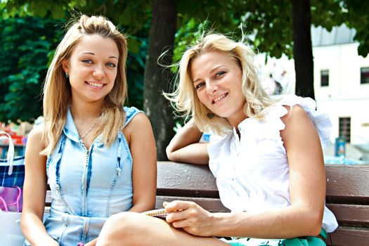 Two beautiful young woman resting on a bench in the park after shopping