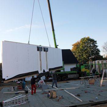 Workmen guiding in a prefabricated wall which is hanging suspended from a crane as it is delivered to a building site for installation onto the cement foundations