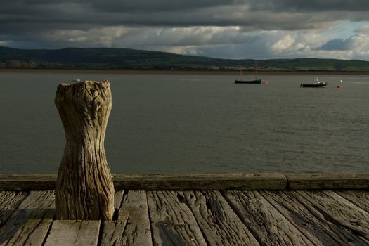 A wooden quay, dock, with an angular, worn, post with the sea and boats under a cloudy sky.