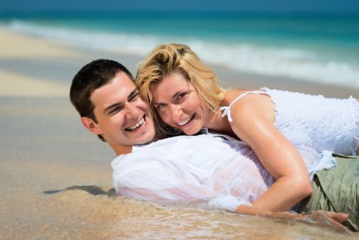 Happy embracing young couple lie on sand of a tropical beach with blue sea on background