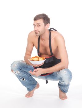 Attractive man trying his hand at baking wearing an apron and carrying a bowl of freshly baked cookies isolated on white