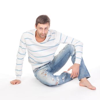 Handsome casual man sitting barefoot in trendy ragged jeans over a white background