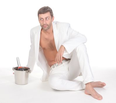 Cool sexy handsome man dressed in a stylish white suit shirtless and barefoot sitting on the floor alongside a bottle of chilled champagne on a white background