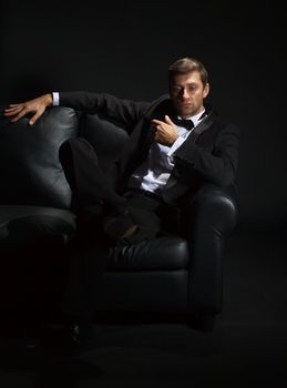 Dramatic portrait of a suave handsome man in a tuxedo and bowtie on couch highlighted in darkness