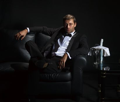 Sexy handsome man in tuxedo sitting in the darkness of a nightclub with an unopened bottle of champagne on ice waiting for his date