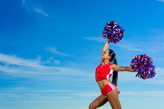 Young cheerleader in red costume with pampon against blue sky