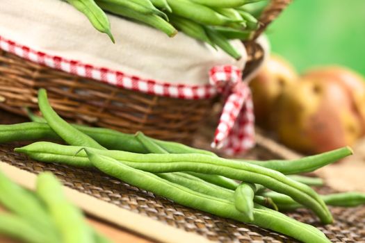 Fresh raw green beans in front of a basket with potatoes in the back (Selective Focus, Focus on the long bean one third into the image )