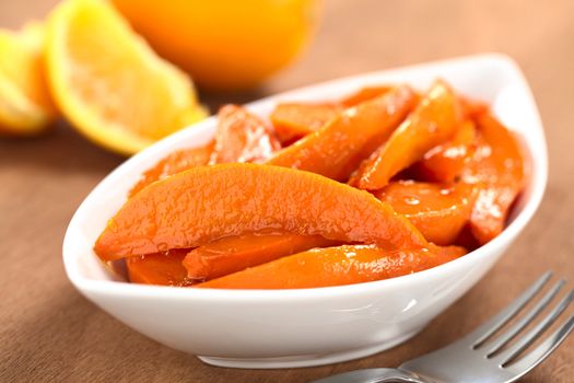 Bowl of sweet potato wedges caramelized with brown sugar and fresh orange juice with a fork on the side and oranges in the back (Selective Focus, Focus on the horizontal wedge in the front)