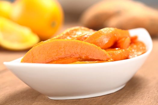 Bowl of sweet potato wedges caramelized with brown sugar and fresh orange juice with sweet potatoes and oranges in the back (Selective Focus, Focus on the horizontal wedge in the front)