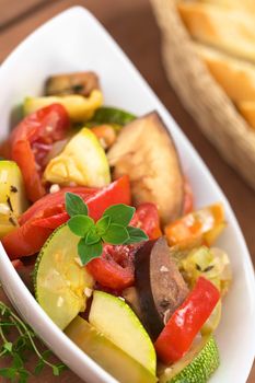 Bowl of fresh homemade Ratatouille made of eggplant, zucchini, bell pepper and tomato and seasoned with herbs (garlic, thyme, oregano) with baguette slices in the back (Selective Focus, Focus on the oregano leaves on the meal)