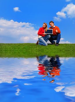 Couple with notebook sitting on grass against sky with reflection on water