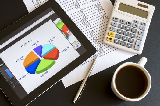 Modern workplace with digital tablet showing charts and diagram on screen, coffee, pen and paper with numbers.