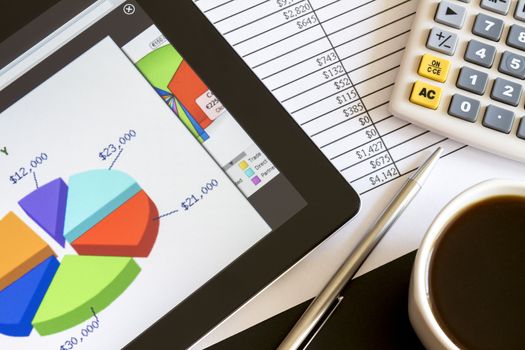 Modern workplace with digital tablet showing charts and diagram on screen, coffee, pen and paper with numbers.