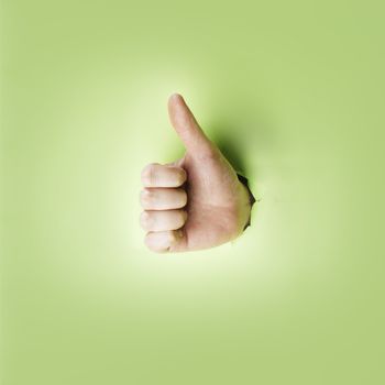Hand making a thumb up gesture through a hole in green paper.