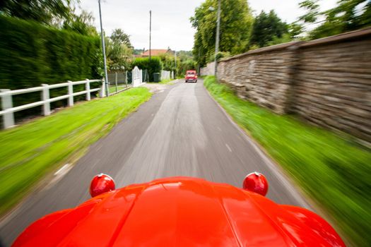Picture taken from inside of a red shiny Citroen 2CV which is driving very fast on a narrow path following another Citroen, on the side green grass, bushes and white fences.