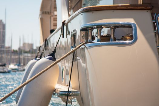 Close-up photo of fenders hanging on the side of a white yacht which is in the port on a sunny day with other ship masts in the background