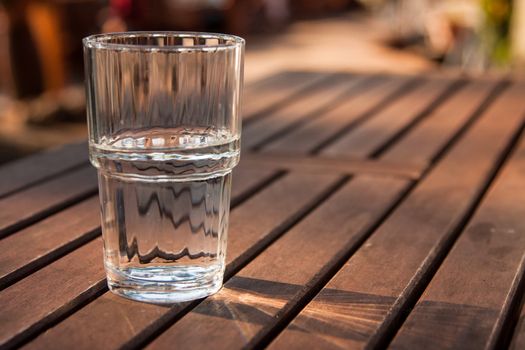 Close-up picture of a glass of water which is half-full standing on a brown wooden table and as the sun shines through the glass the reflection can be seen on the table
