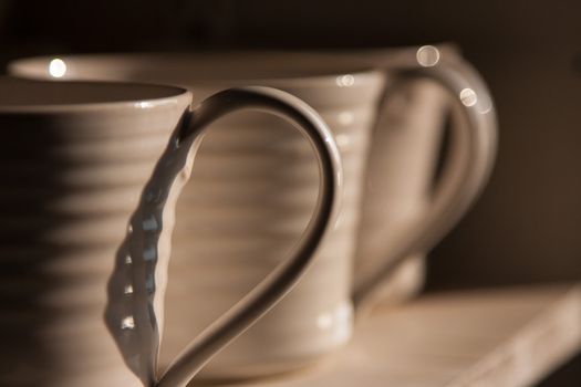 Close-up photo of the handles of white mugs in half-shadow on a shelf
