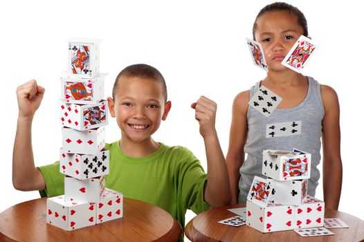 A young boy smiles happily when he wins a card building competition against his siter, whose tower collapses.