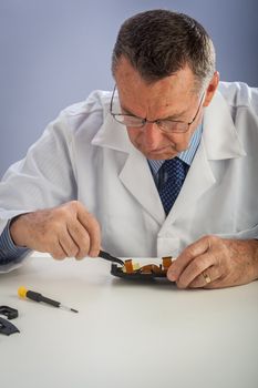 An older male wearing a white lab coat and repairing electronic equipments, like a technician or a repair man.