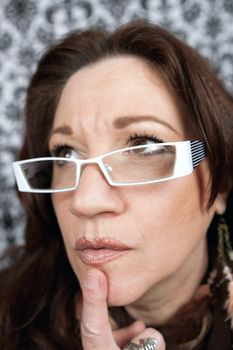 Brunette middle aged woman wearing white framed glasses with her finger on her chin in deep contemplation.  Shallow depth of field.