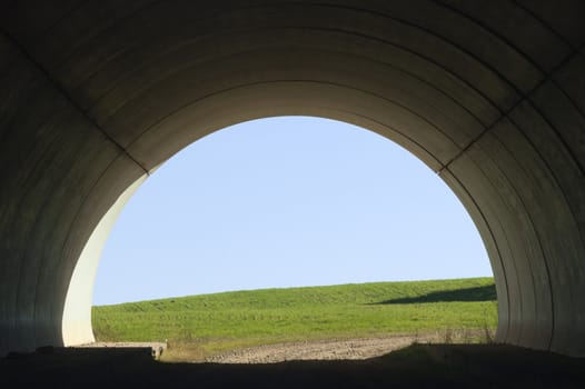 Dirt road under a concrete tunnel of a highway overlooking a green field
