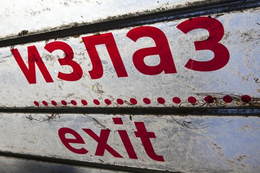 A worn out exit sign written in red letters in english and cyrilic ("izlaz") against a white background with black stipes.