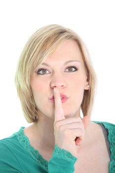 Attractive young blonde woman requesting quiet with a hushing gesture raising her finger to her lips isolated on white