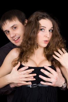 Portrait of playful man holding pretty woman by her breast. Isolated on black