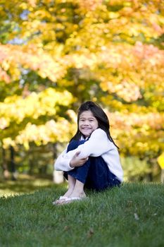 Little girl hugging knees sitting on lawn against bright autumn leaves in background