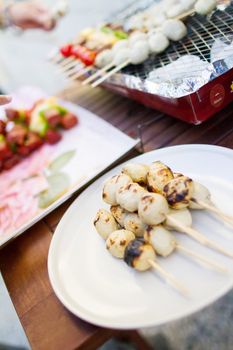 Thai style BBQ chicken balls on skewer grilling on a plate in outdoor cooking area.