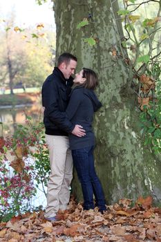 Passionate young couple standing close together looking into each others eyes in front of a tree trunk on a lakeshore