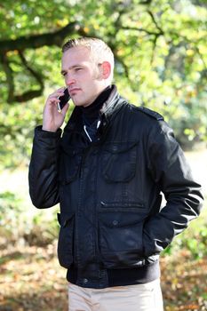 Handsome trendy young man standing talking on his mobile phone outdoors in autumn woodland Handsome trenduy young man standing talking on his mobile phone outdoors in autumn woodland