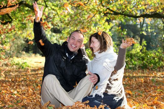 Happy attractive young couple sitting close togather on the ground in woodland while out on a date playing with autumn leaves