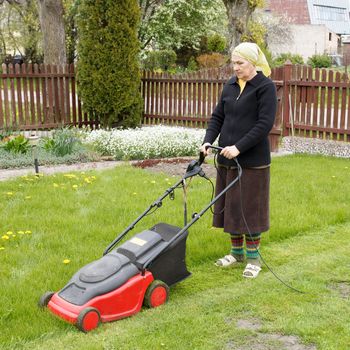 senior woman mowing grass with electric mower in his flower garden