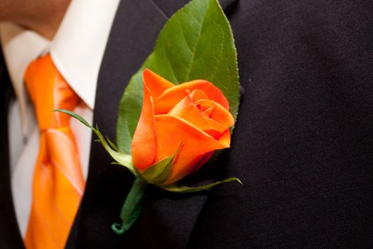 A groom's orange boutineer upon his black tuxedo jacket at a wedding with orange roses.