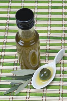 A bottle of olive oil  and a china spoon with olives on a bamboo mat