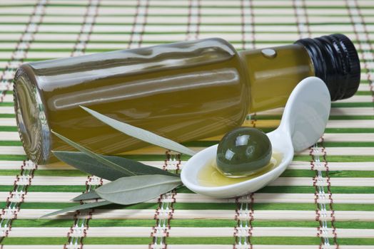 A bottle of olive oil  and a china spoon with olives on a bamboo mat