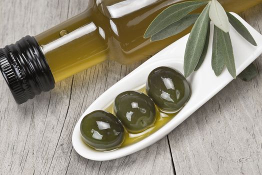 Olive oil bottle and some olives in a china spoon on a wooden surface