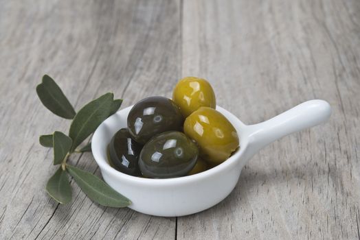 A china spoon with olives and leaves on a wooden surface
